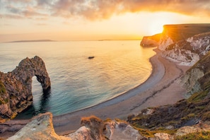 Paisaje costero con una formación rocosa destacada, una costa curva y una puesta de sol.