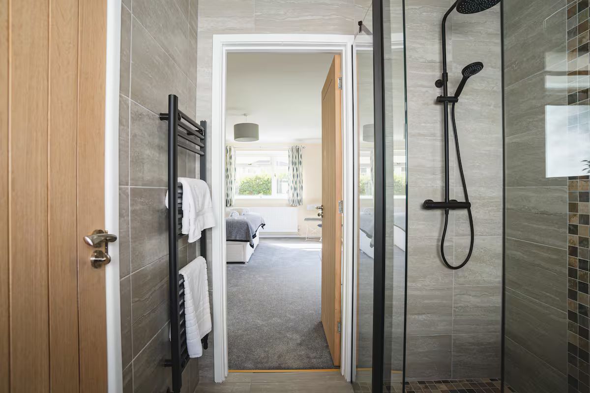 Ein modernes Badezimmer mit einer Duschkabine aus Glas, einem Handtuchhalter mit weißen Handtüchern und einem sichtbaren Schlafzimmer mit Bett und Fenster.