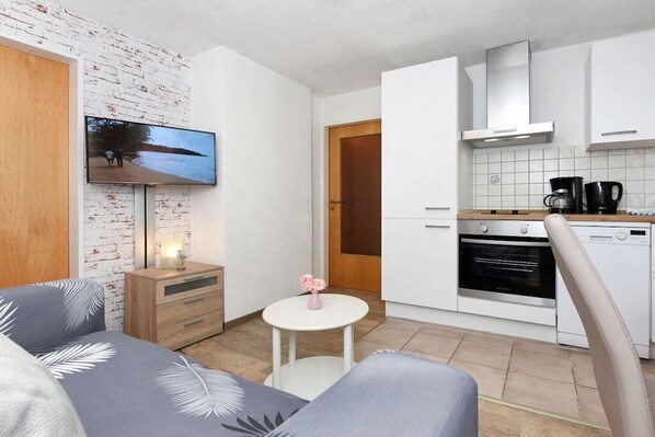 Een knus woongedeelte met een flatscreen televisie aan een muur met baksteenmotief, een kitchenette met moderne apparatuur en een klein, rond tafeltje met een vaas.