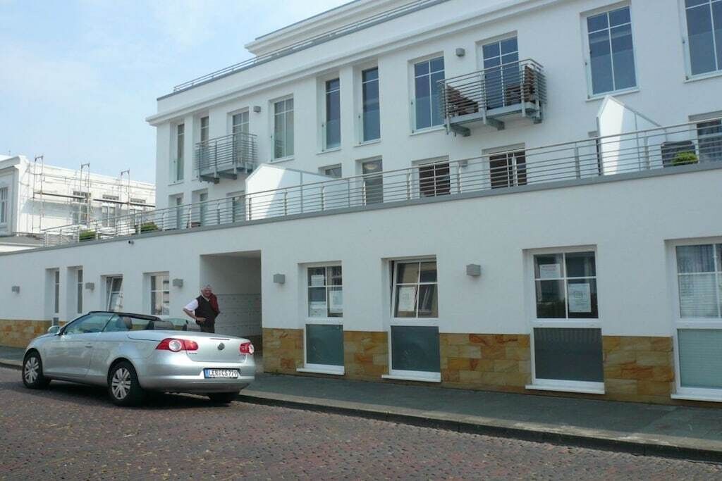 Ein mehrstöckiges Gebäude mit weißer Fassade, ein geparktes Auto und eine Person in der Nähe des Eingangs.