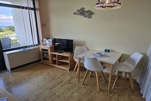 Ein Zimmer mit Holzboden, einem Fernseher auf einem Holzbodenständer, einem Tisch mit vier Stühlen und einem Fenster mit Jalousien.