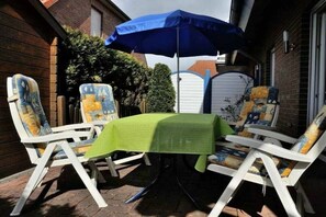 Een terras met een tafel, afgedekt met een groene tafelkleed, twee witte stoelen met gekleurde kussens, en een blauwe parasol voor de schaduw.