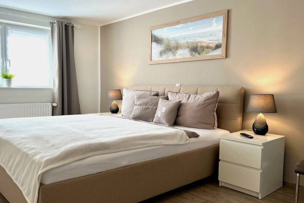 Una camera da letto con un letto, lampade sui comodini, un comodino, una finestra con tende e un quadro appeso al muro.