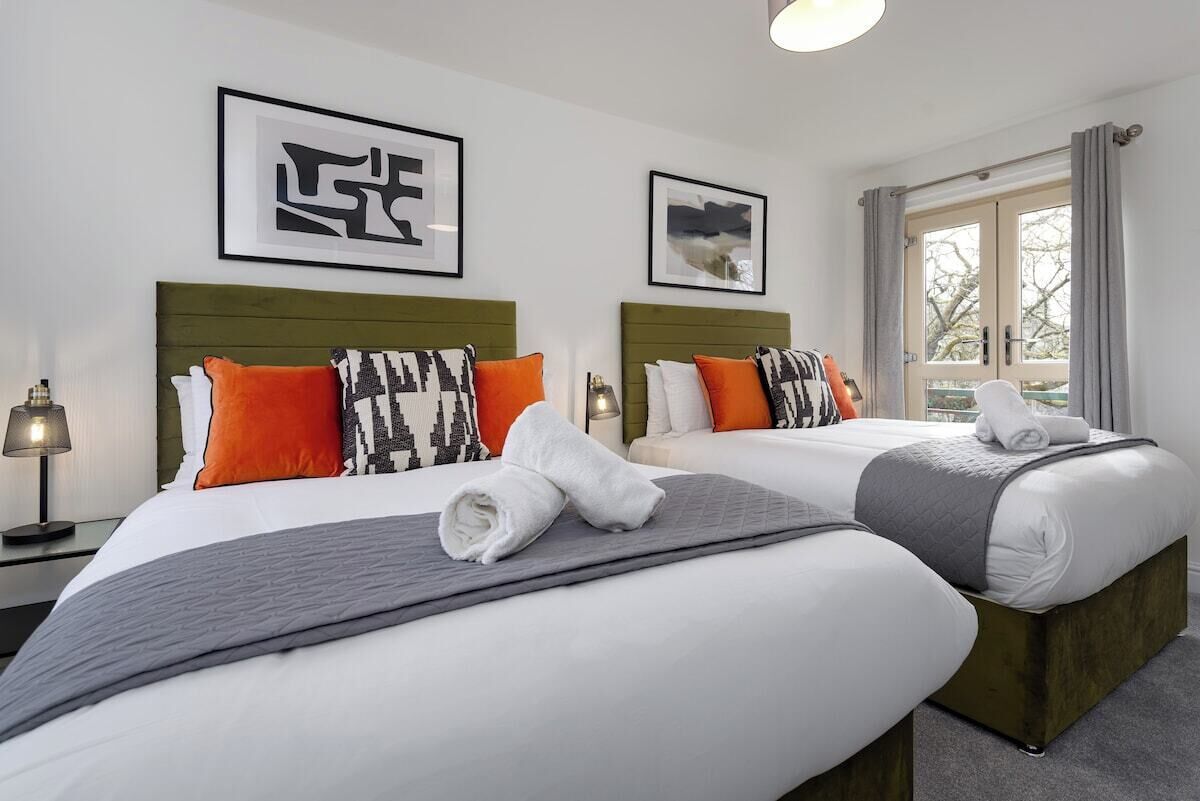 A hotel room with two beds, each with a green headboard, white bedding, and patterned pillows. There are framed artworks on the walls and a window with curtains.