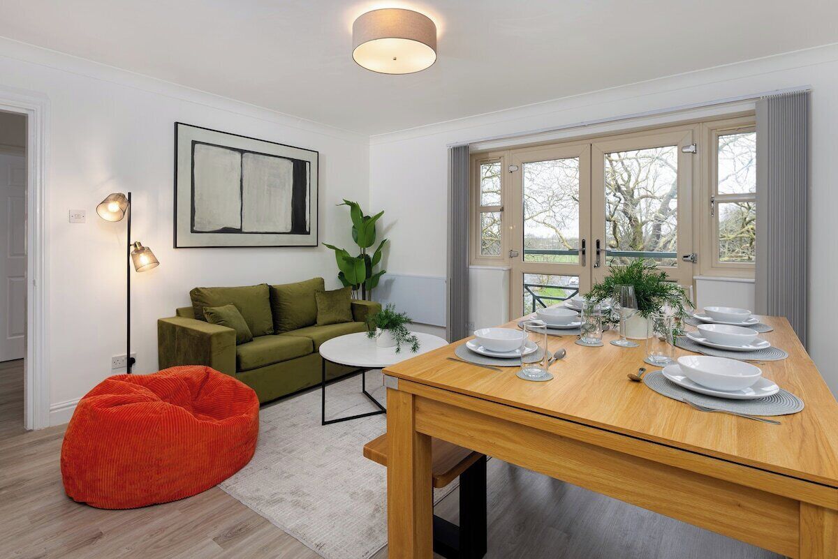 A dining area with a wooden table set for four, a green sofa, a red bean bag, and a floor lamp.