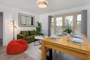 A dining area with a wooden table set for four, a green sofa, a red bean bag, and a floor lamp. - Flatzy - Peaceful Sefton Park Luxury Apartment (Liverpool)