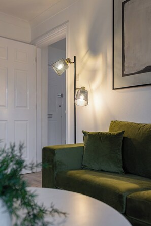 Ein Zimmer mit einem grünen Sofa, zwei Lampen, einer weißen Tür und einem gerahmten Kunstwerk an der Wand.