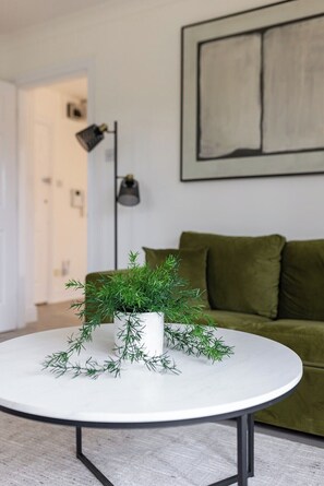 Ein runder, weißer Tisch mit einem Blumentopf in der Mitte, ein grünes Sofa im Hintergrund und eine Stehlampe links.