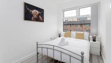 Quarto com estrutura de cama de metal, colcha branca, toalha, mesa de cabeceira com abajur e janela com vista para edifícios.