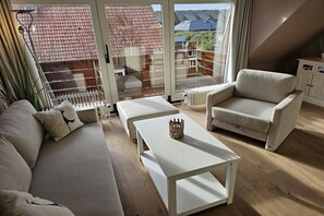 A living room with a sofa, armchair, and coffee table, a radiator, and a view through a glass door. - Holiday apartment with 1 bedroom (Wenningstedt-Braderup (Sylt))