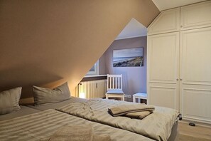 Ein Schlafzimmer mit schrägem Dach, einem Bett mit gestreifter Bettwäsche, einem Sessel, einem kleinen Tisch und einem Kleiderschrank.