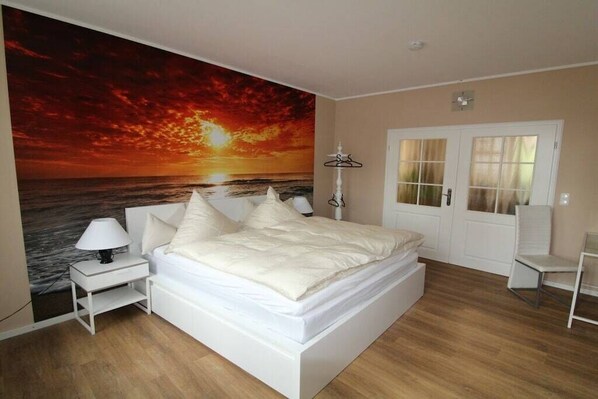 A bedroom with a bed, a nightstand, a lamp, a chair, a door, and a wall with a sunset painting.
