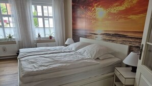 Ein Schlafzimmer mit einem Bett, Nachttischlampen, einem Fenster mit Vorhängen und einem Wandbild mit Sonnenuntergang.