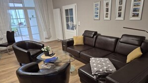 Ein modernes Wohnzimmer mit einer Ledersofa, einem Glastisch und einem Sessel. An der Wand hängen gerahmte Bilder und ein Übergang zur einem Balkon.