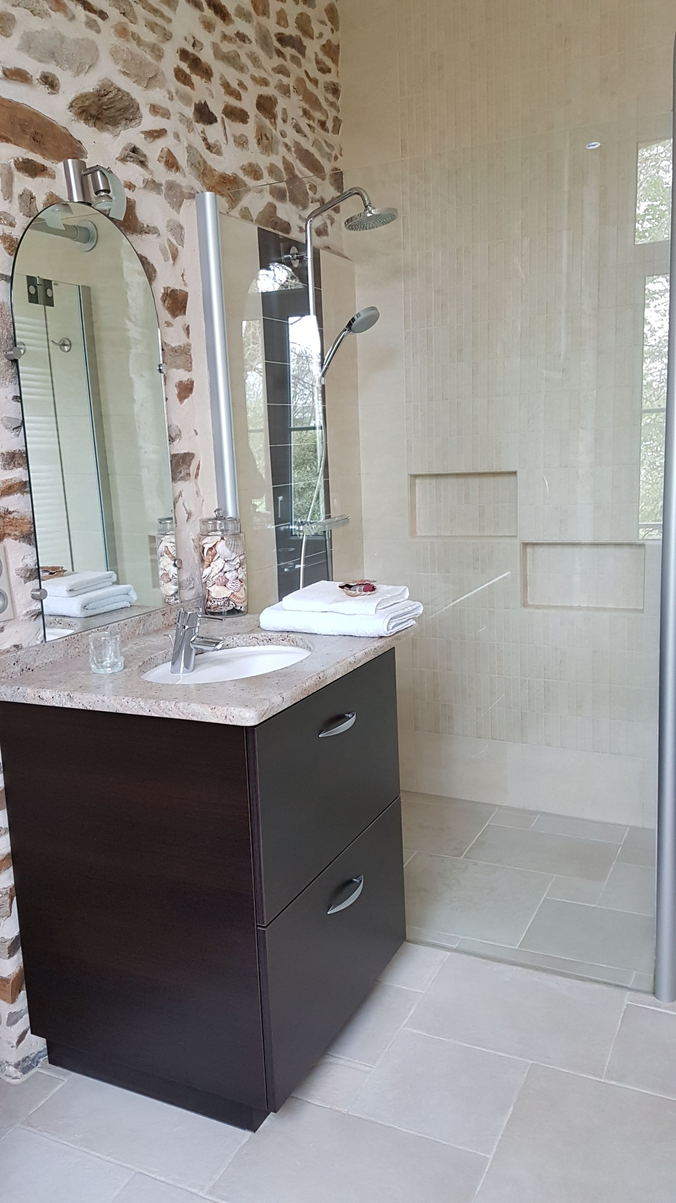 Een moderne badkamer met een stenen muur, een grote spiegel, een wastafel met een glas en een douchehoek.