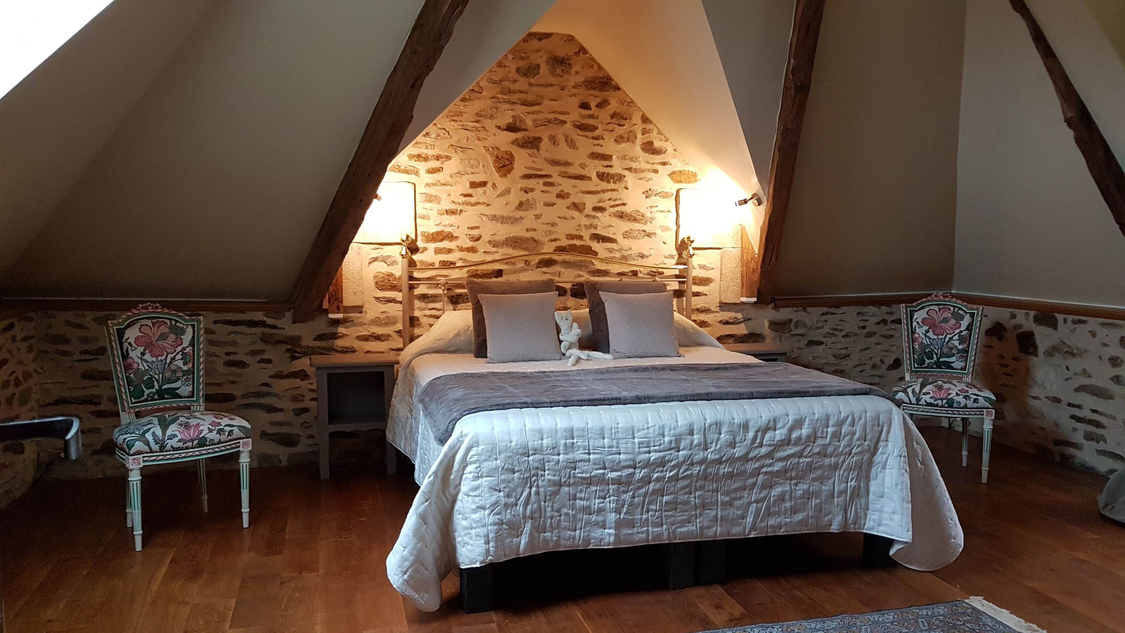 A bedroom with a stone wall, a bed with pillows, two chairs, and a nightstand.