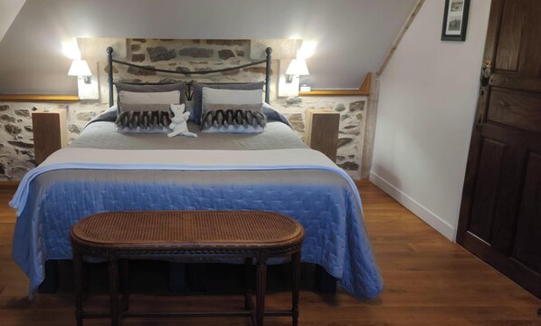 A bedroom with a stone wall, a bed with a blue bedspread, two bedside lamps, and a wicker chair. - The Manor of Bigotière (Pleslin-Trigavou)