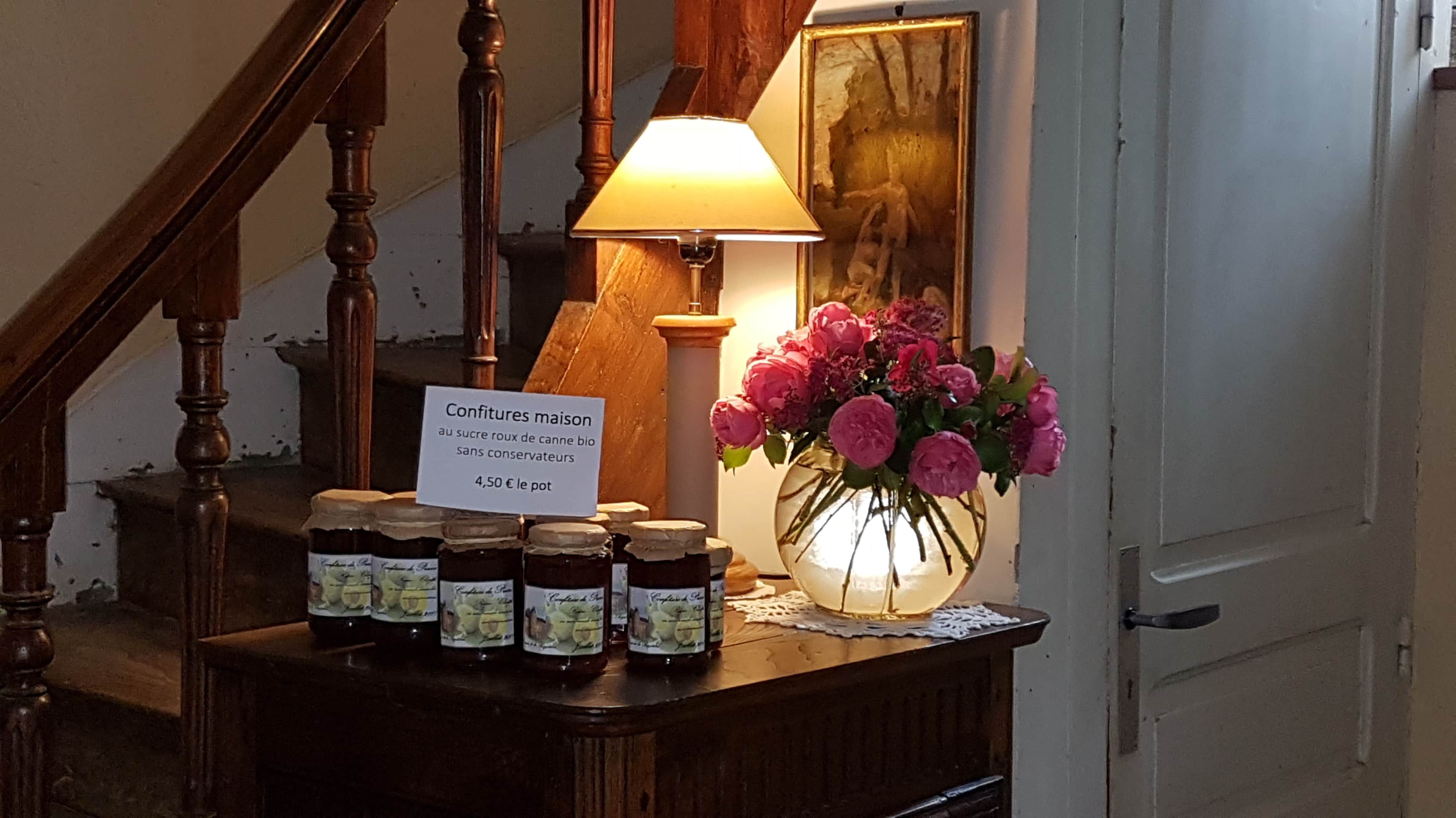 A wooden counter with jars of jam, a sign indicating the price, a vase of flowers, and a lamp.