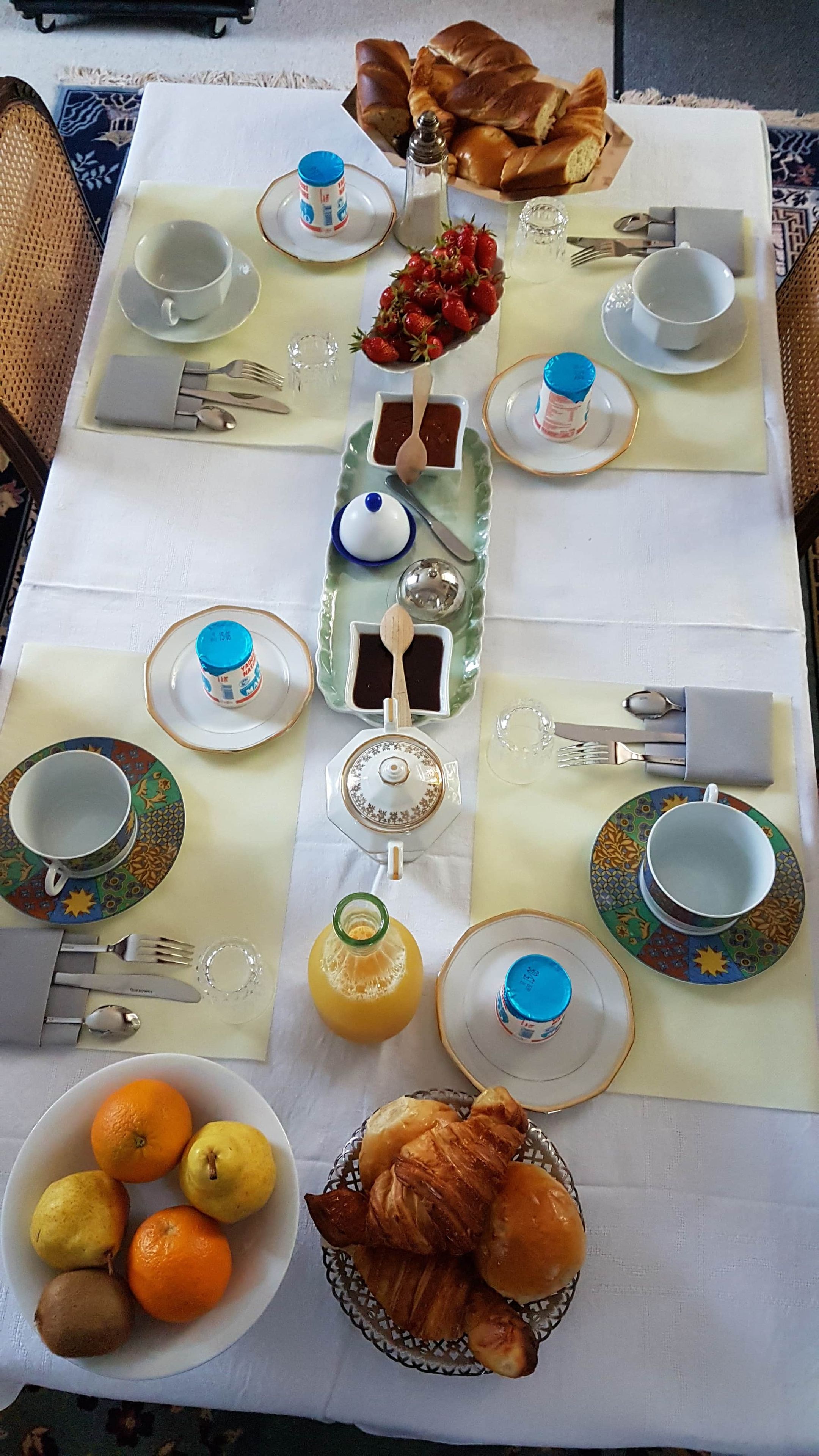Ein Frühstückstisch gedeckt mit Tellern, Besteck, einer Teekanne, einem Orangensaftkrug und einer Obstschale.