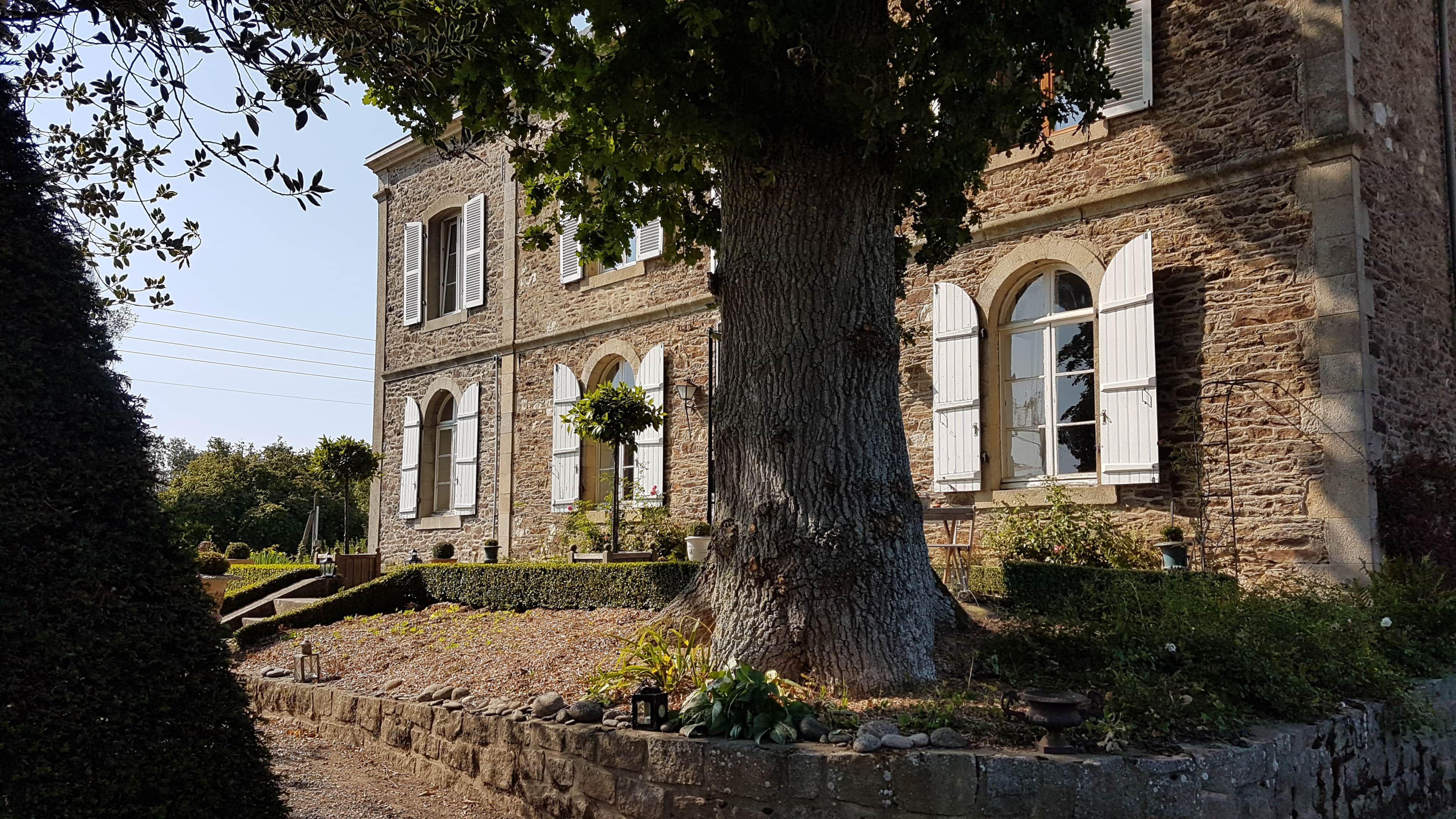 Un bâtiment en pierre, doté de fenêtres en arc de cercle et de volets, entouré d’un mur de pierre et d’un grand arbre.