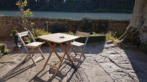 An outdoor table and chairs set on a stone patio with a view of a body of water and trees in the background. - The Manor of Bigotière (Pleslin-Trigavou)