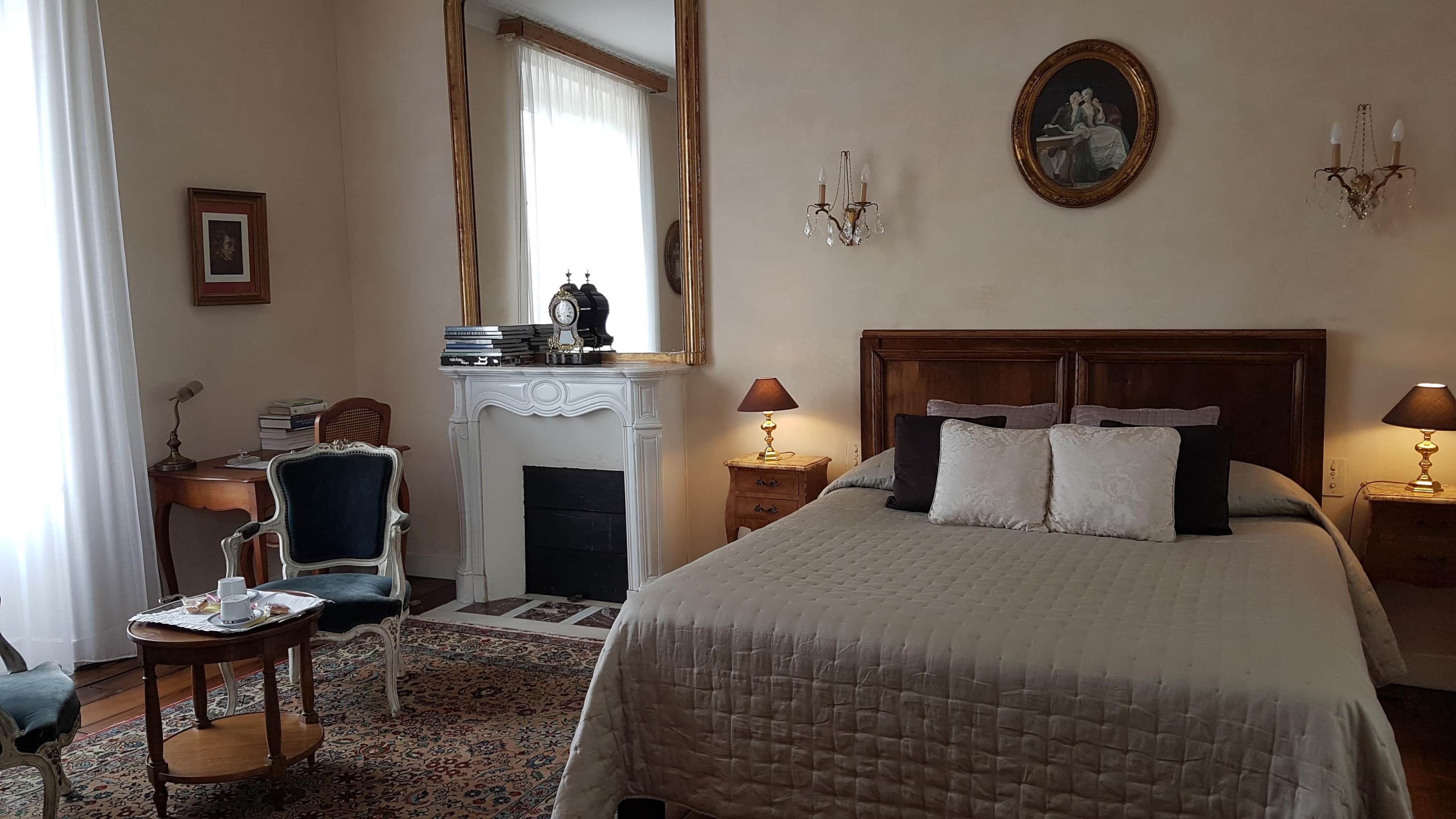 Une chambre d’hôtel avec un grand lit, un coin salon comprenant un fauteuil et une petite table, une cheminée et un miroir.