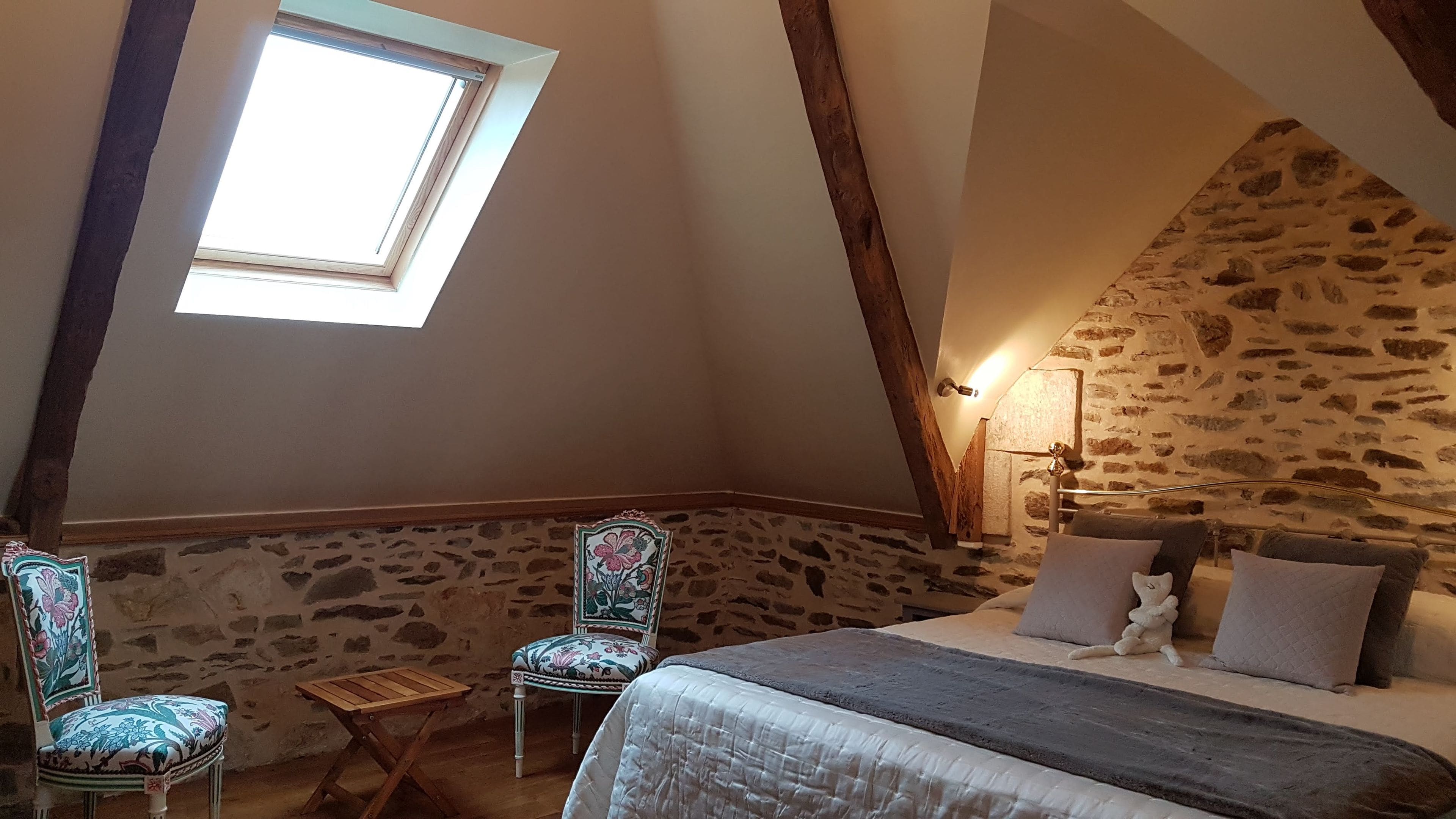 Un dormitorio con pared de piedra inclinada, tragaluces, cama con almohadas, una silla y una pequeña mesa.