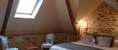 Un dormitorio con pared de piedra inclinada, tragaluces, cama con almohadas, una silla y una pequeña mesa.