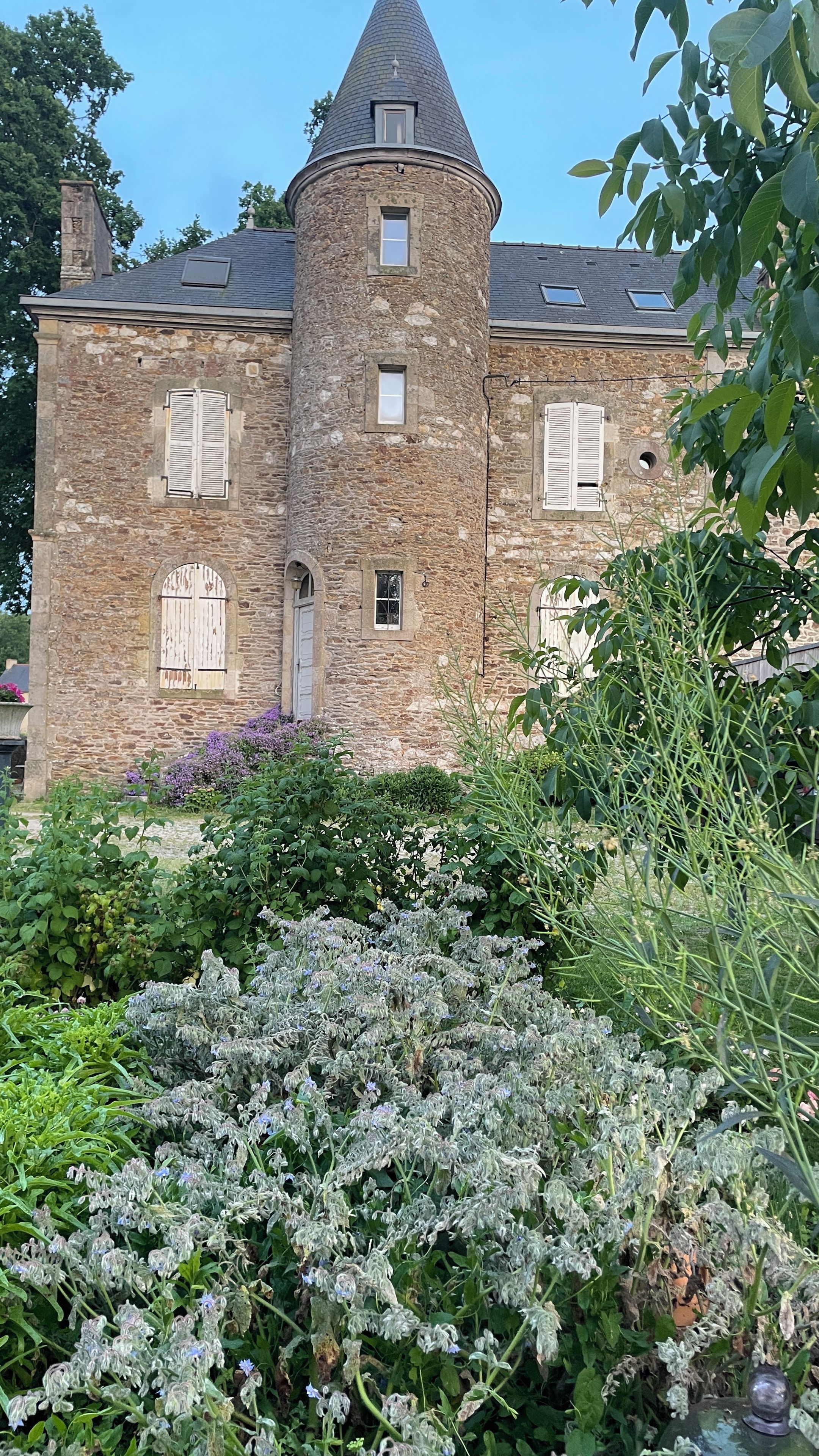A stone building with a conical roof, multiple windows with shutters, and a garden with various plants.