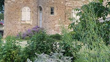 A stone building with a conical roof, multiple windows with shutters, and a garden with various plants.