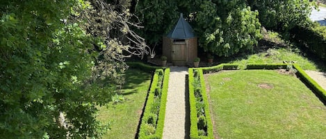 Ein formeller Garten mit einem zentralen Weg, der zu einem kleinen Gebäude führt, umgeben von ordentlich geschnittenen Hecken und Blumentöpfen.