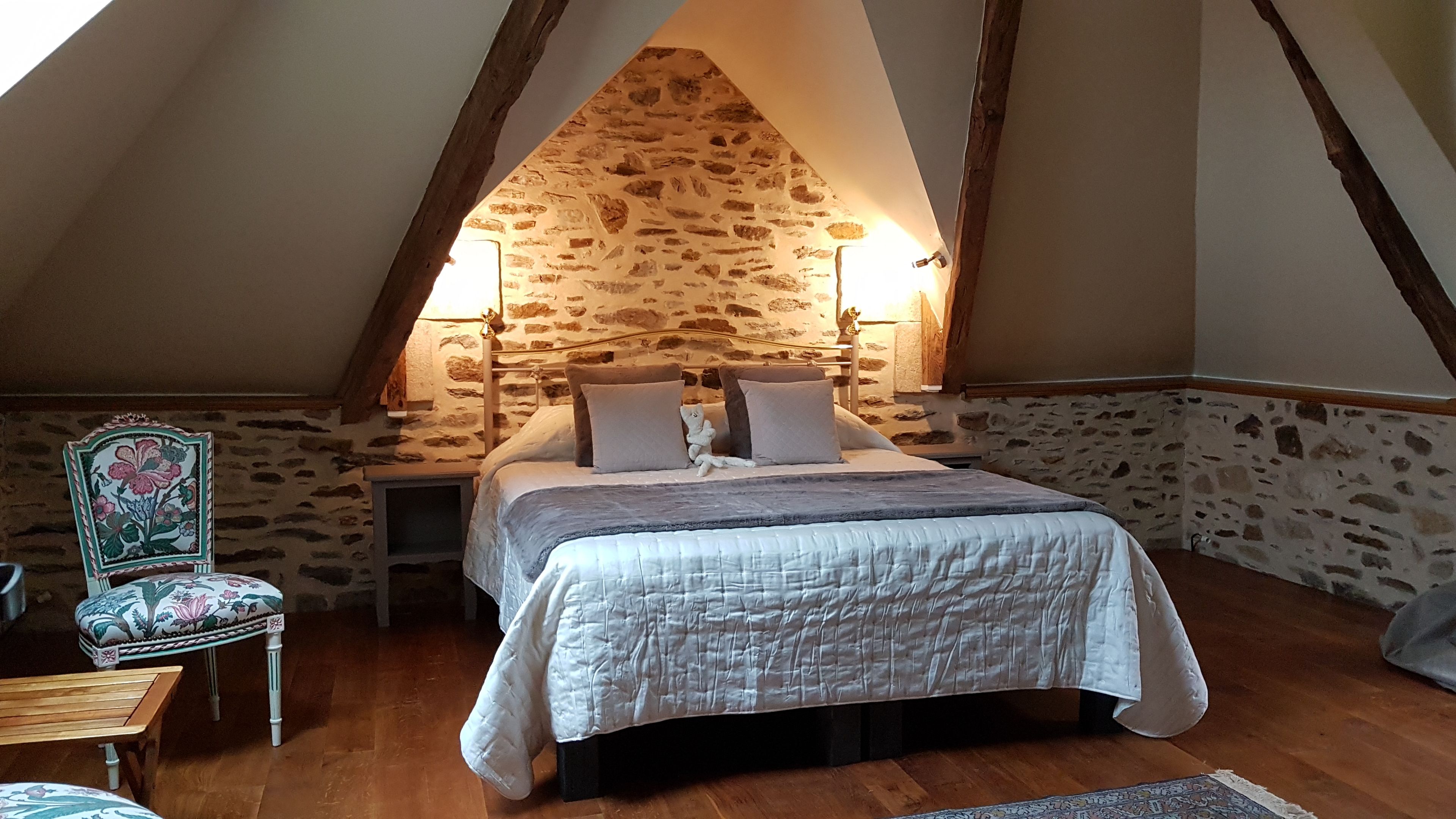 Quarto com parede de pedra, cama com almofadas, mesa de cabeceira, cadeira com tecido floral e piso de madeira.