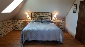 A bedroom with a stone wall, a sloped ceiling, a bed with a headboard, two bedside lamps, and a wooden door. - The Manoir de la Bigotière—Jean-Sébastien Bach Room (Pleslin-Trigavou)