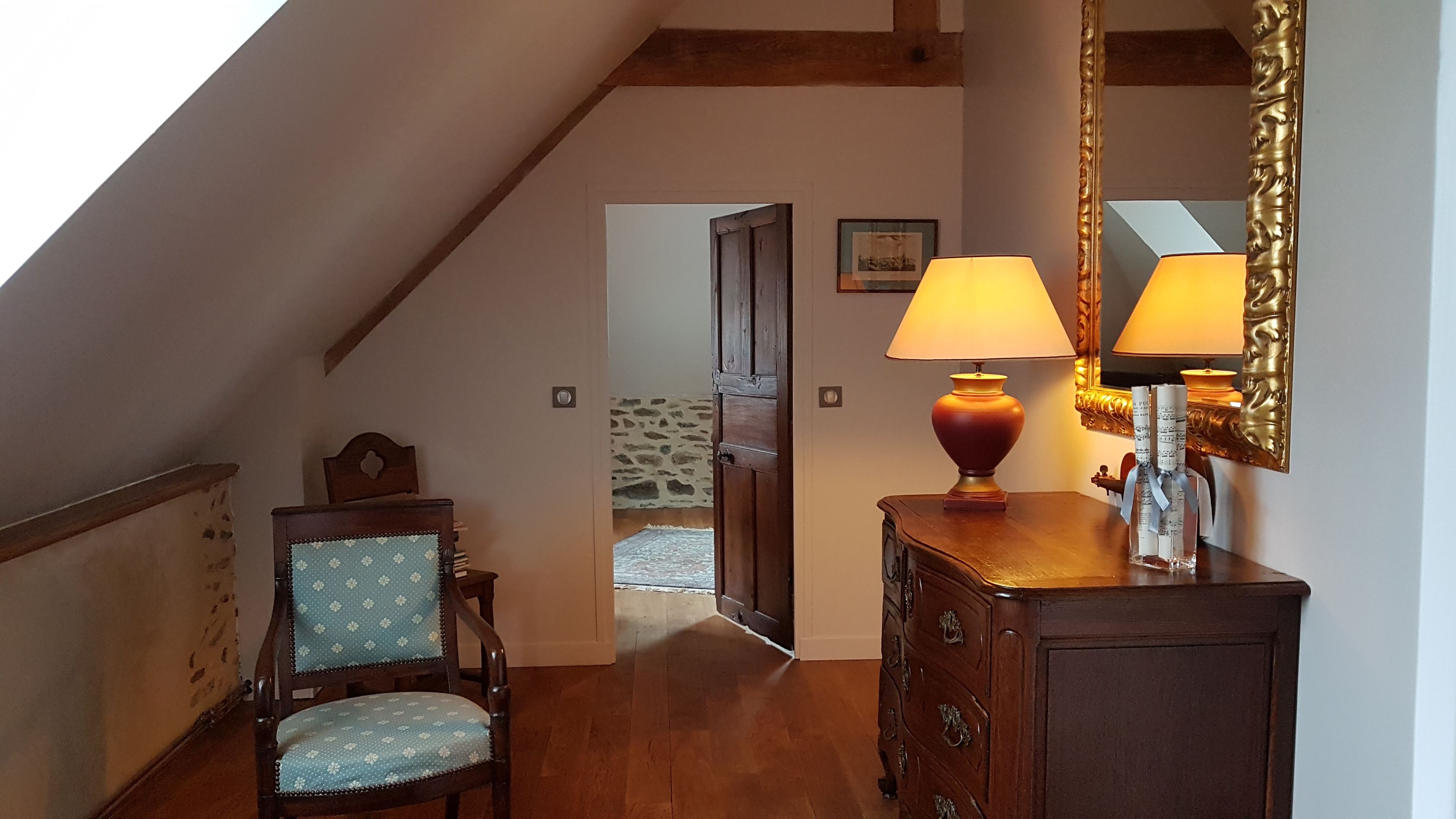 Ein Zimmer mit einem Holzbett, einer Holzkommode, einer Lampe und einem Spiegel. Eine Tür führt in einen anderen Raum.