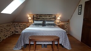 Een slaapkamer met een schuine kap, stenen muren, een bed met grijze beddengoed, twee bedlampjes en een houten bankje.