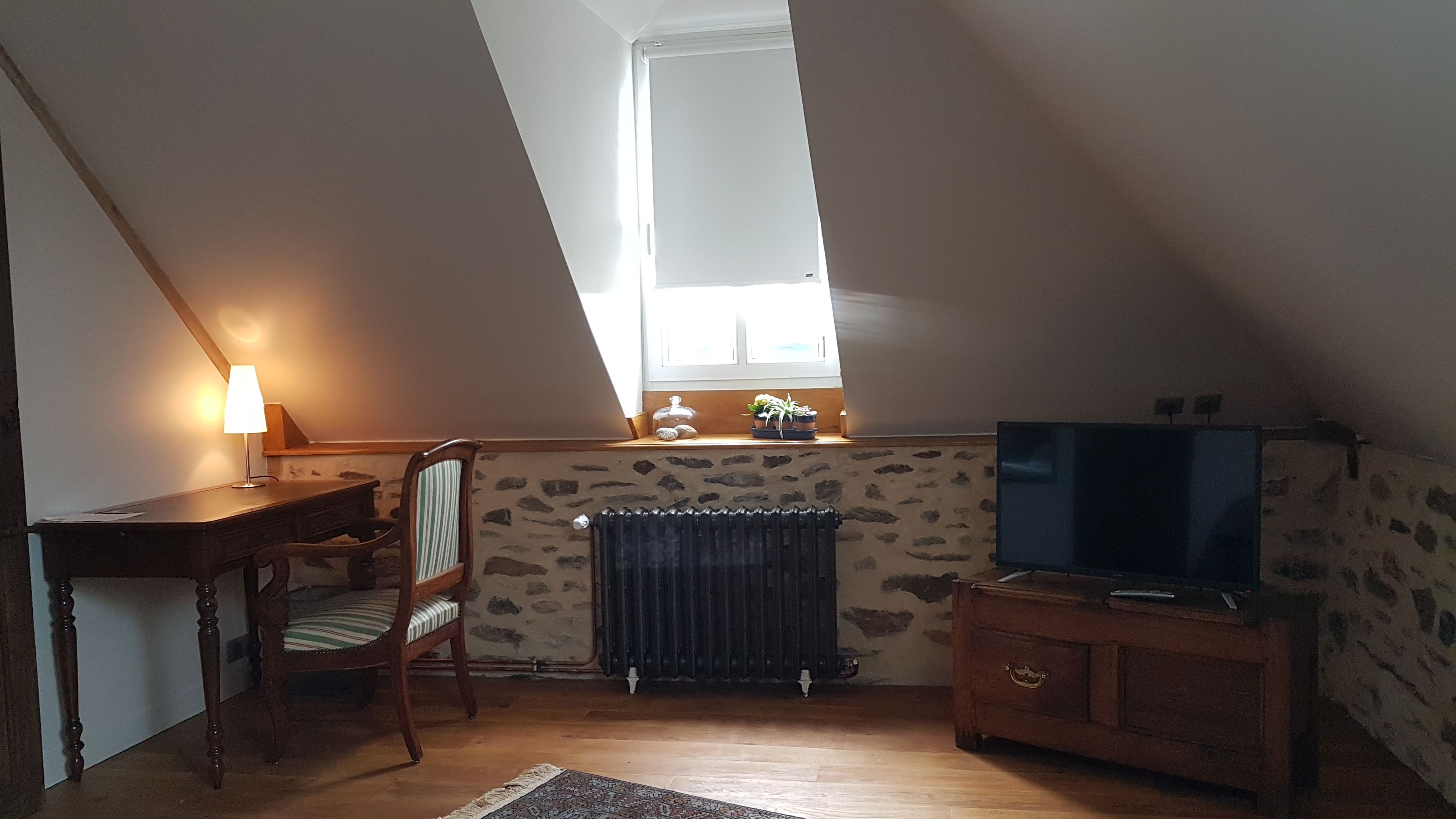 A cozy attic room with a sloped ceiling, a wooden desk with a lamp, a chair, a radiator, and a television on a wooden cabinet.