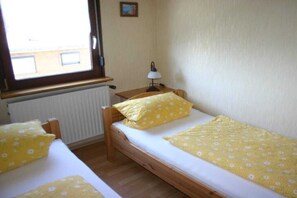 Ein Zimmer mit zwei Einzelbetten, einem Fenster, einem Heizkörper und einem kleinen Tisch mit einer Lampe.