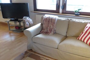 Ein Wohnzimmer mit einer Couch, einem Fernseher, einem kleinen Tisch mit Dekoration und einem Fenster mit Blick.
