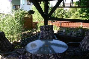 Eine Terrasse im Freien mit einem runden Glastisch und gemusterten Stühlen, umgeben von Grünpflanzen und einem Holzzaun.