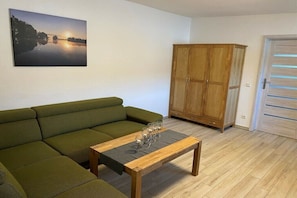 Ein Wohnzimmer mit einem grünen Sofa, einem Couchtisch aus Holz, einem Holzkabinett und einer Tür mit horizontalen Jalousien.