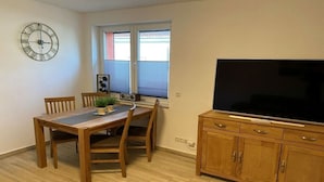 A dining area with a wooden table and chairs, a clock on the wall, a window with blinds, and a TV on a wooden cabinet. - Holiday home in Zachow with swimming pool (Zachow)
