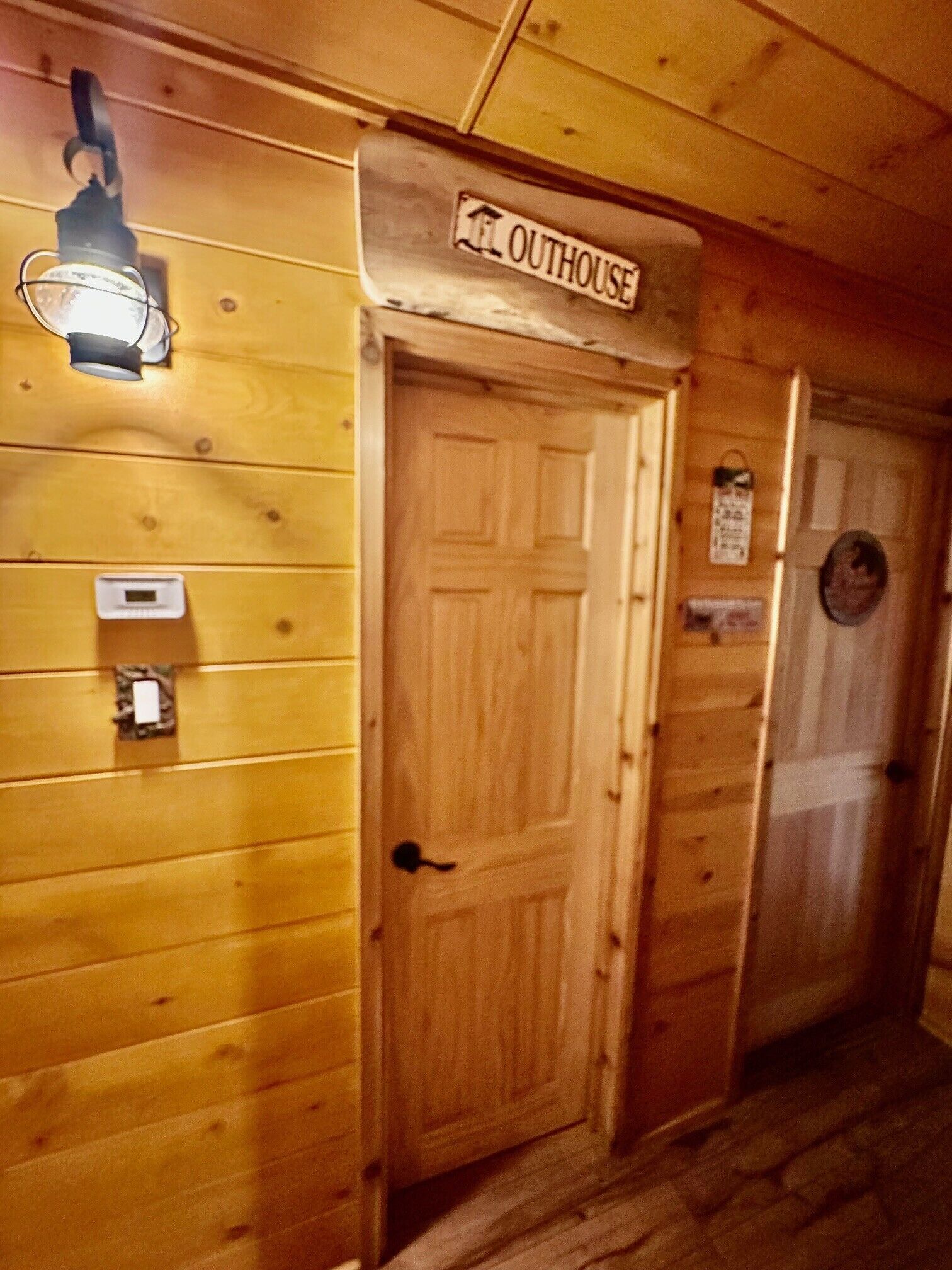 Um corredor de madeira com uma porta identificada como ‘OUTHOUSE’ e uma luminária fixada na parede.