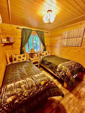 A cabin-style room with two beds, a window with curtains, a lamp, and a wall-mounted shelf with books. - A Custom-Built Swiss-Style Luxury Mountain Retreat - Lower Level 2 BR Unit (Mount Jackson)
