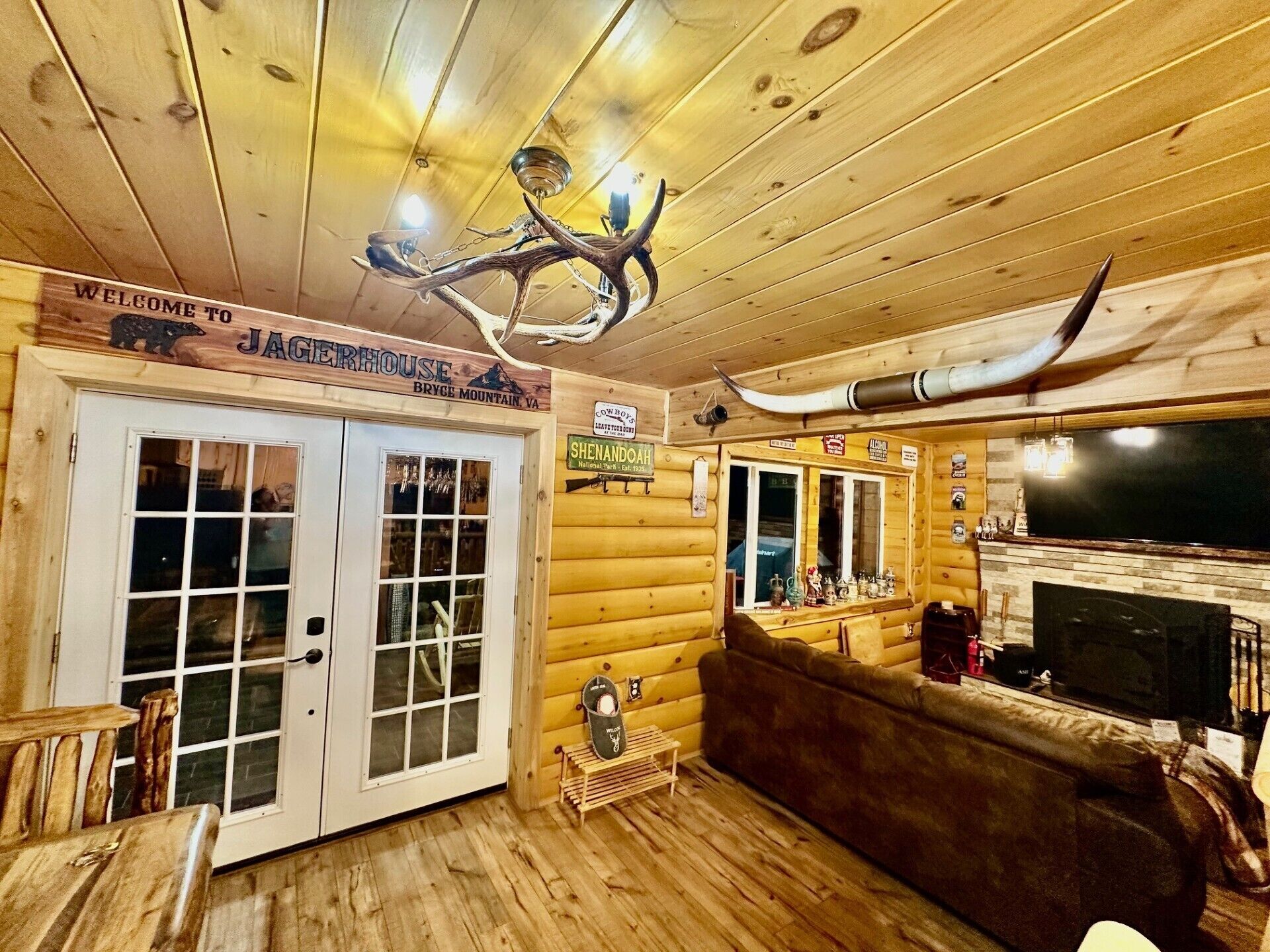 A log cabin interior with a wooden ceiling, a mounted antler chandelier, a fireplace, and a sign reading 'Welcome to Jagerhouse.'