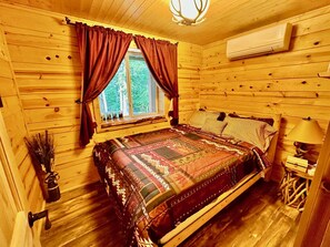 A wooden cabin bedroom with a large bed, a window with curtains, a ceiling light, and an air conditioning unit.