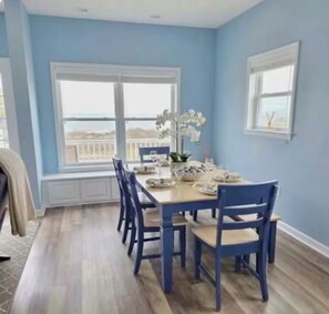 A dining area with a table set for four, blue chairs, and a view through a large window. - Bayside Ortley Retreat 6 Badges And Parking (Seaside Heights)