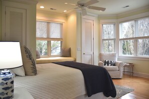 A bedroom with a bed, a chair, a window seat, and a ceiling fan.