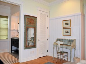A room with a white door, a mirror, a small table with a chair, and a window with blinds.