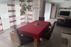 Un comedor con mantel rojo, sillas, una planta y un televisor al fondo.