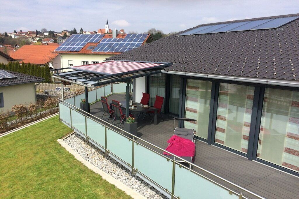 Een modern huis met een glazen terras, zonnepanelen op het dak, en uitzicht over de wijk.